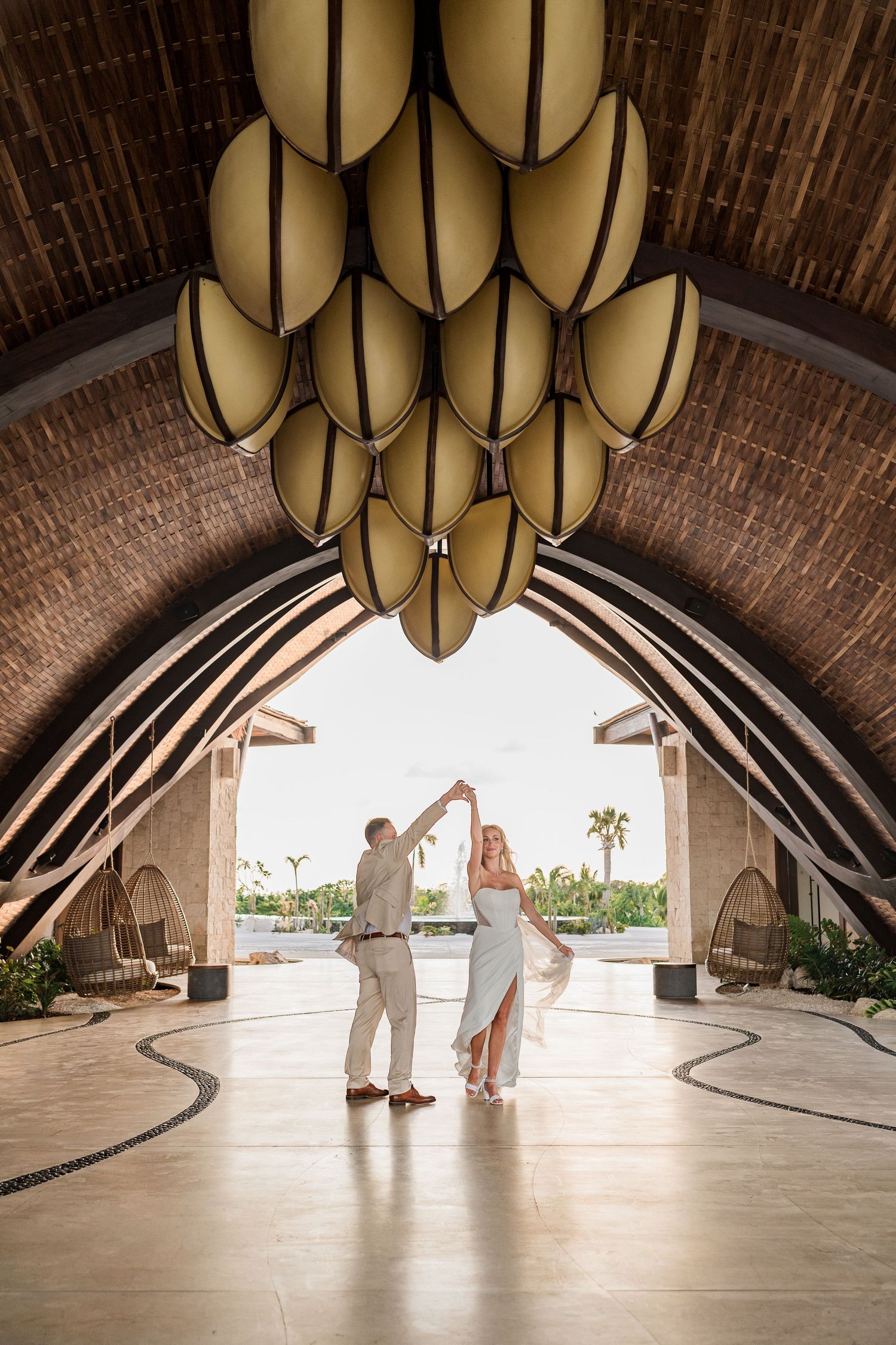 Bride and groom dance elegantly beneath a stunning oval pendant chandelier in an arched tropical venue.