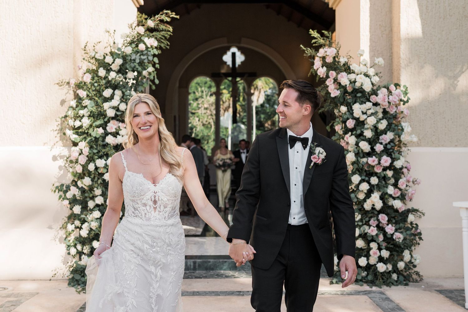 Bride and groom holding hands smiling under a floral arch with pink and white flowers at an elegant wedding venue.