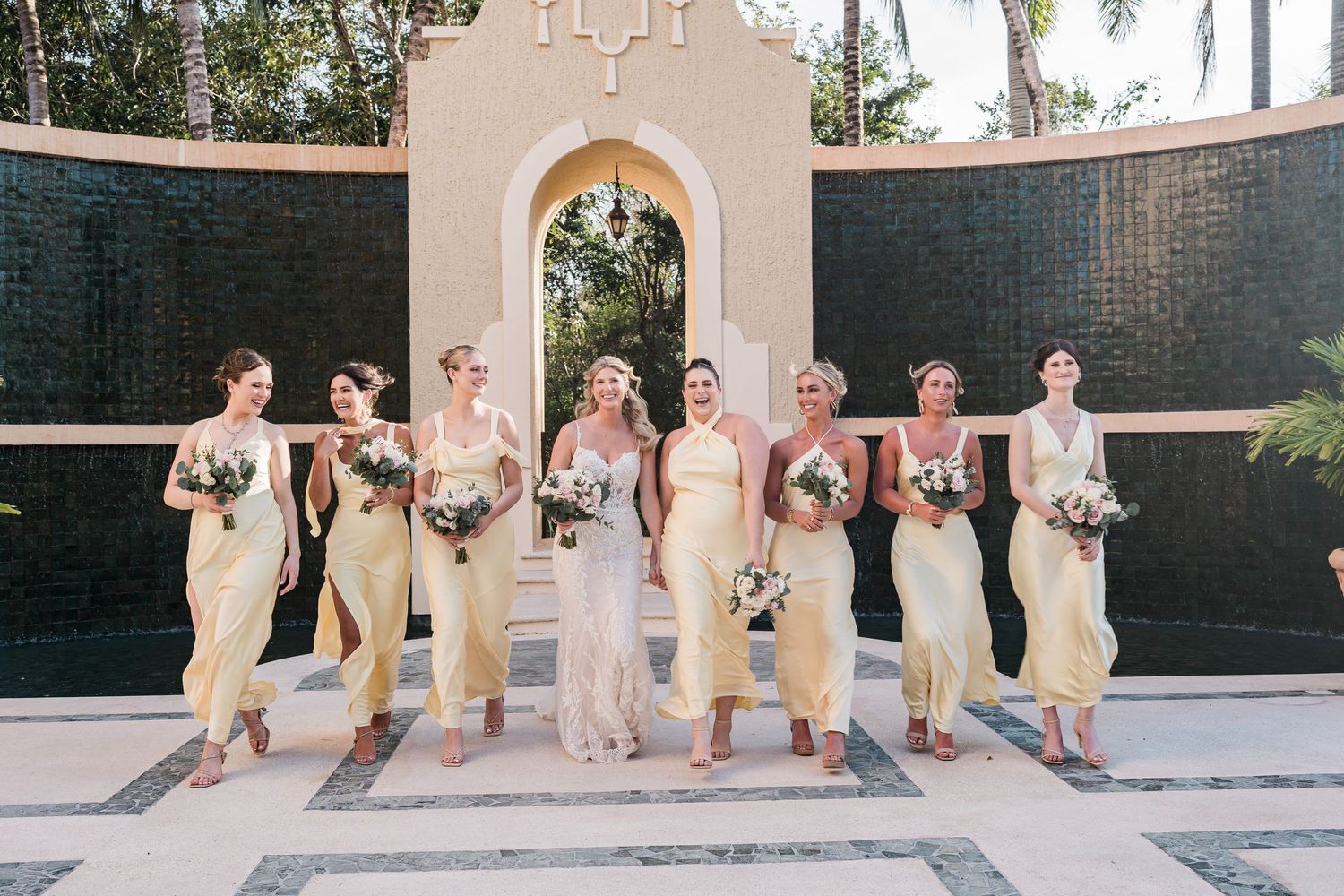 Bride and bridesmaids in yellow dresses walking together outdoors at a tropical wedding venue.