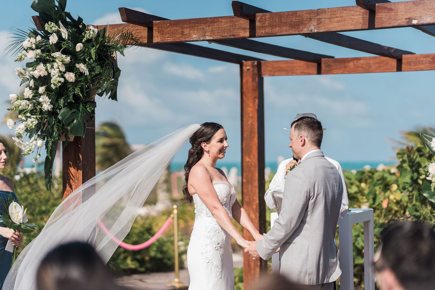 Bride and groom exchange vows at outdoor beach wedding ceremony under wooden pergola with white floral arch.