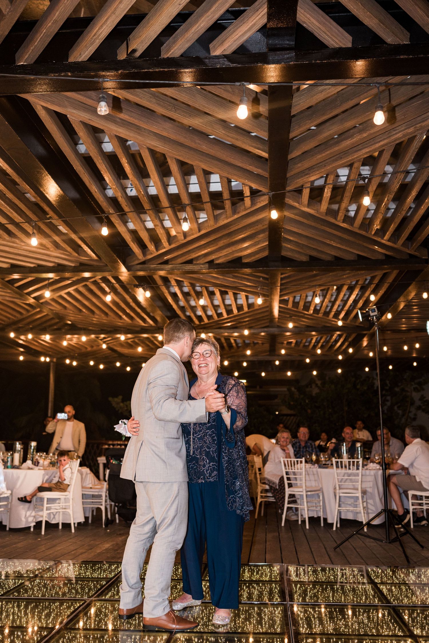 Groom dancing with mother under string lights at rustic outdoor wedding reception with white chairs.