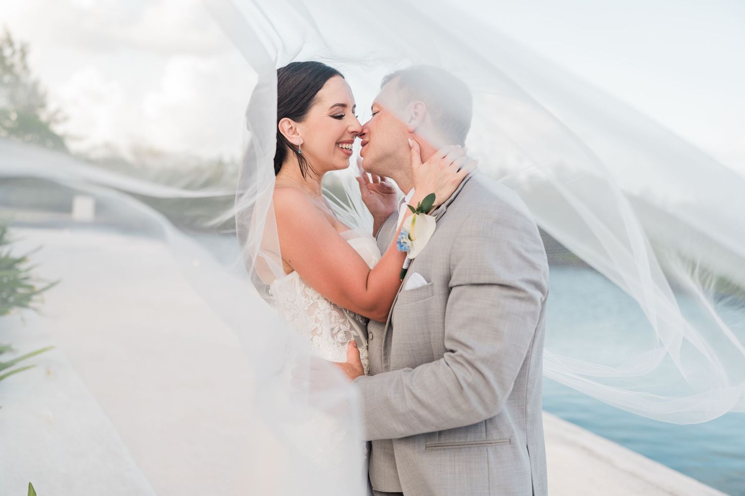 Bride and groom share a romantic moment under flowing veil at outdoor waterfront wedding ceremony.