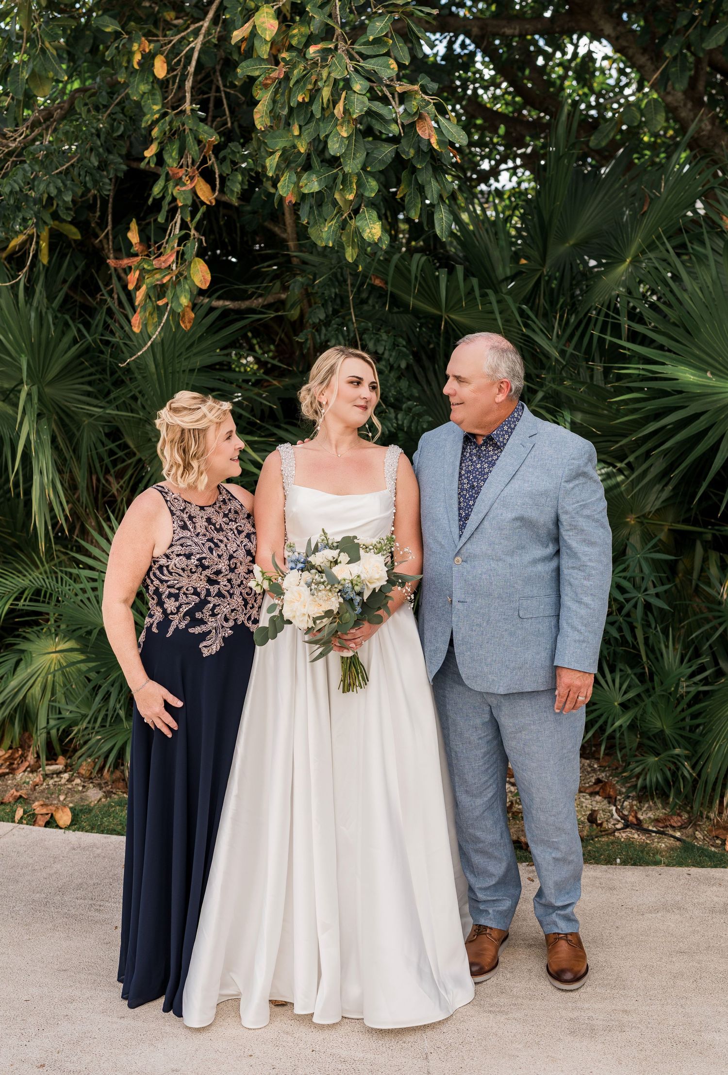 Bride in white gown holds bouquet flanked by parents in formal attire outdoors with tropical greenery.