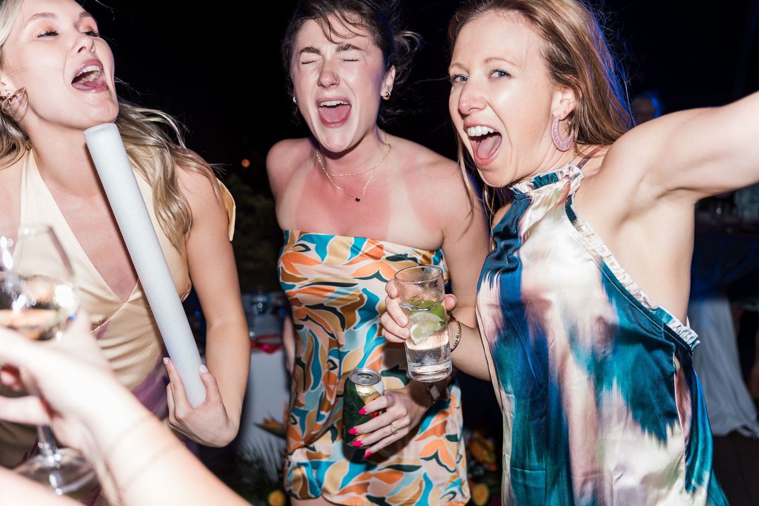 Three women laughing and dancing at a party, holding drinks and wearing colorful dresses at a nighttime event.