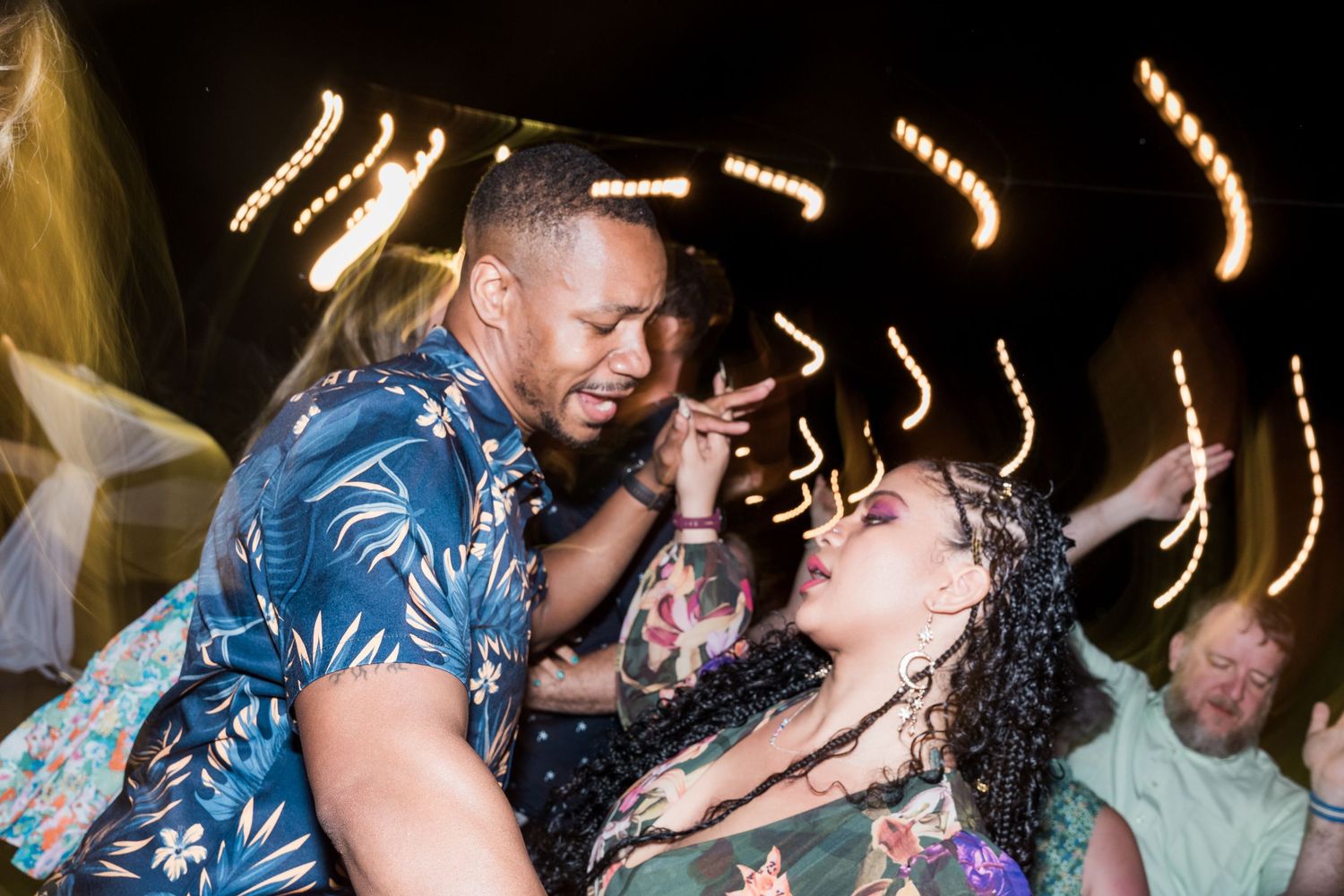 Couple dancing closely at an outdoor night party with bokeh string lights glowing in the background.