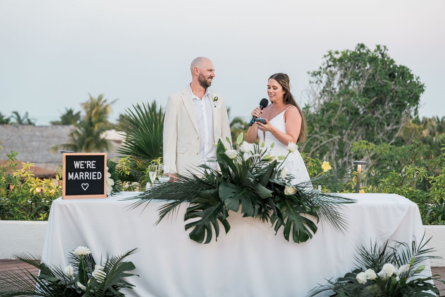 Newlywed couple stands at decorated sweetheart table with We're Married sign, tropical greenery, and white flowers at outdoor wedding.