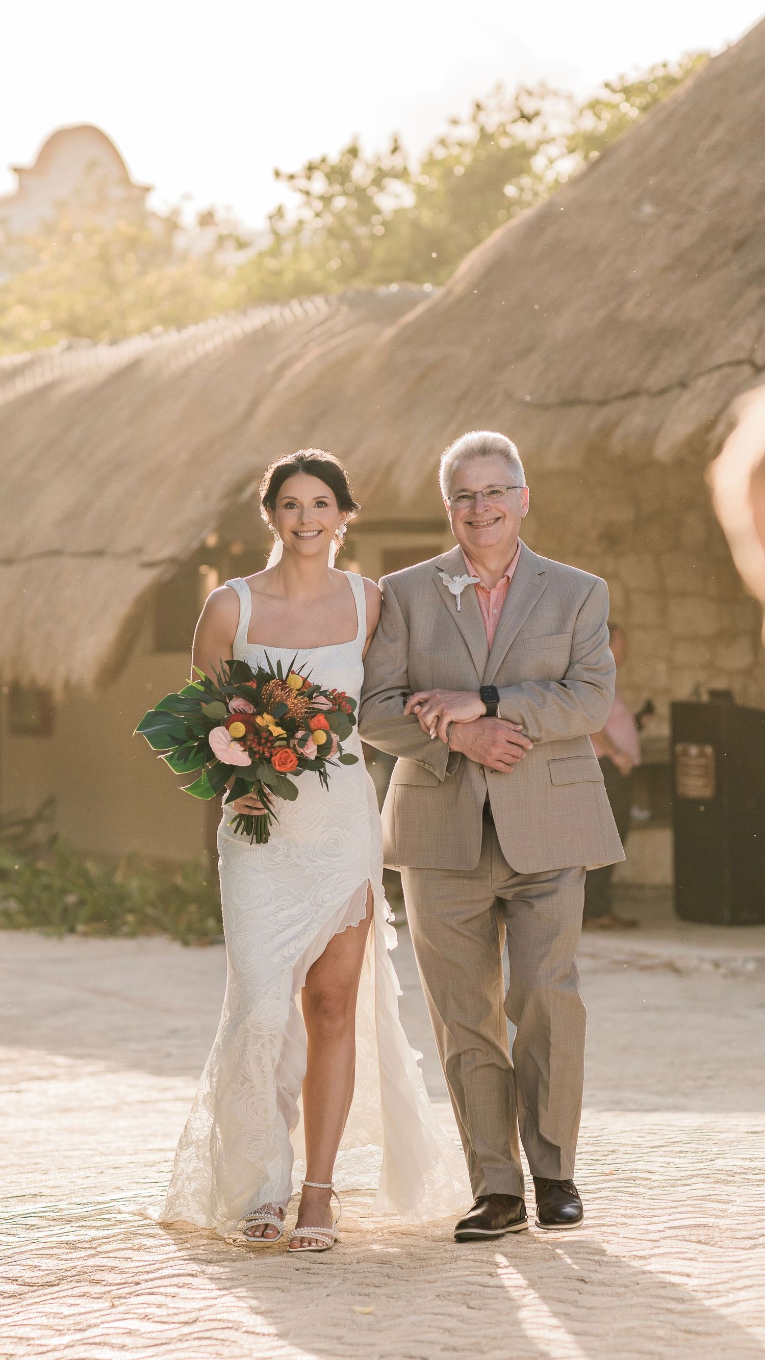 Bride in white lace gown with tropical bouquet walks arm-in-arm with father in golden sunset light.