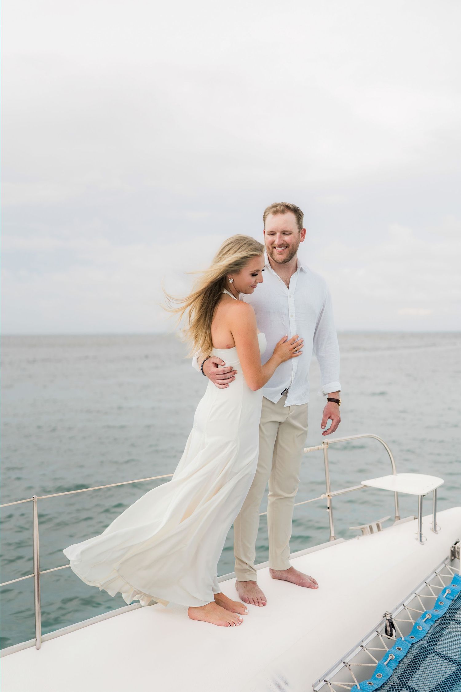 Cancun Engagement Photoshoot - Couple embracing on yacht bow in white outfits, ocean backdrop, woman in flowing dress, man in linen shirt.