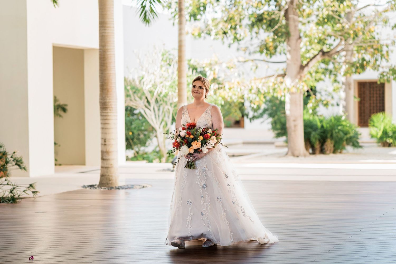 Secrets Akumal Wedding - Bride in a floral ball gown holding a vibrant bouquet walks through an elegant outdoor venue with palm trees.