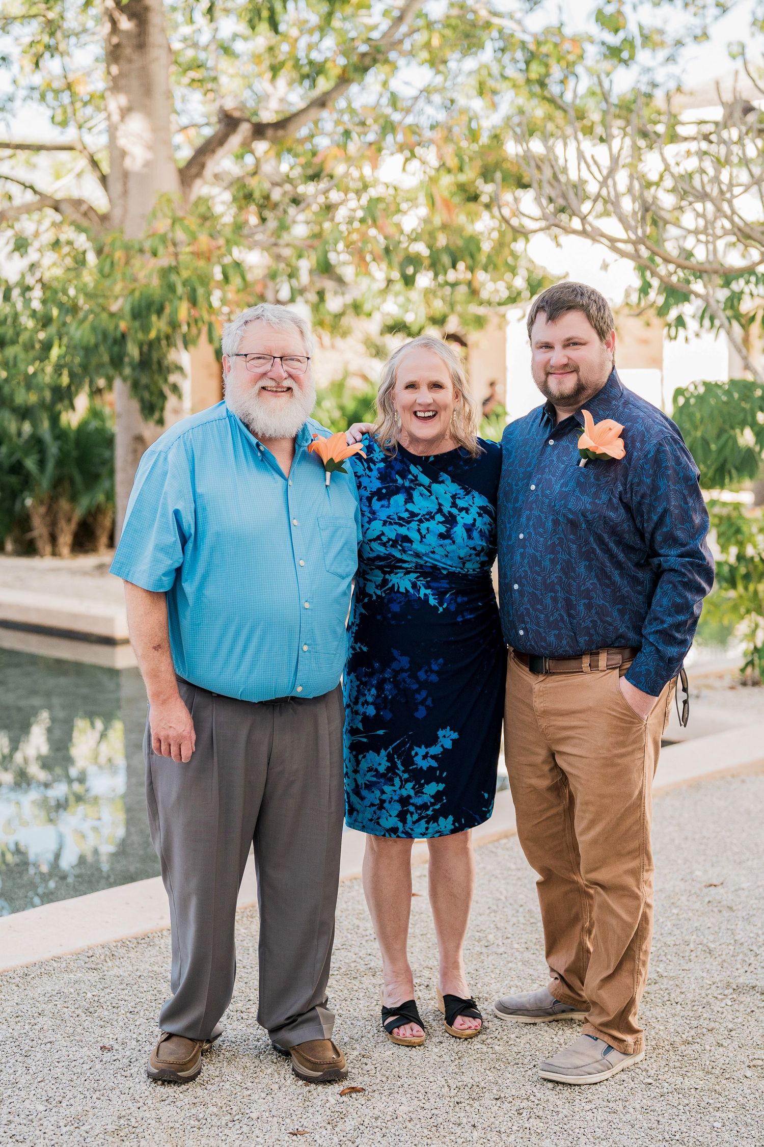 Secrets Akumal Wedding -Three smiling people in blue outfits pose outdoors near a pool, wearing orange floral accessories at a formal event