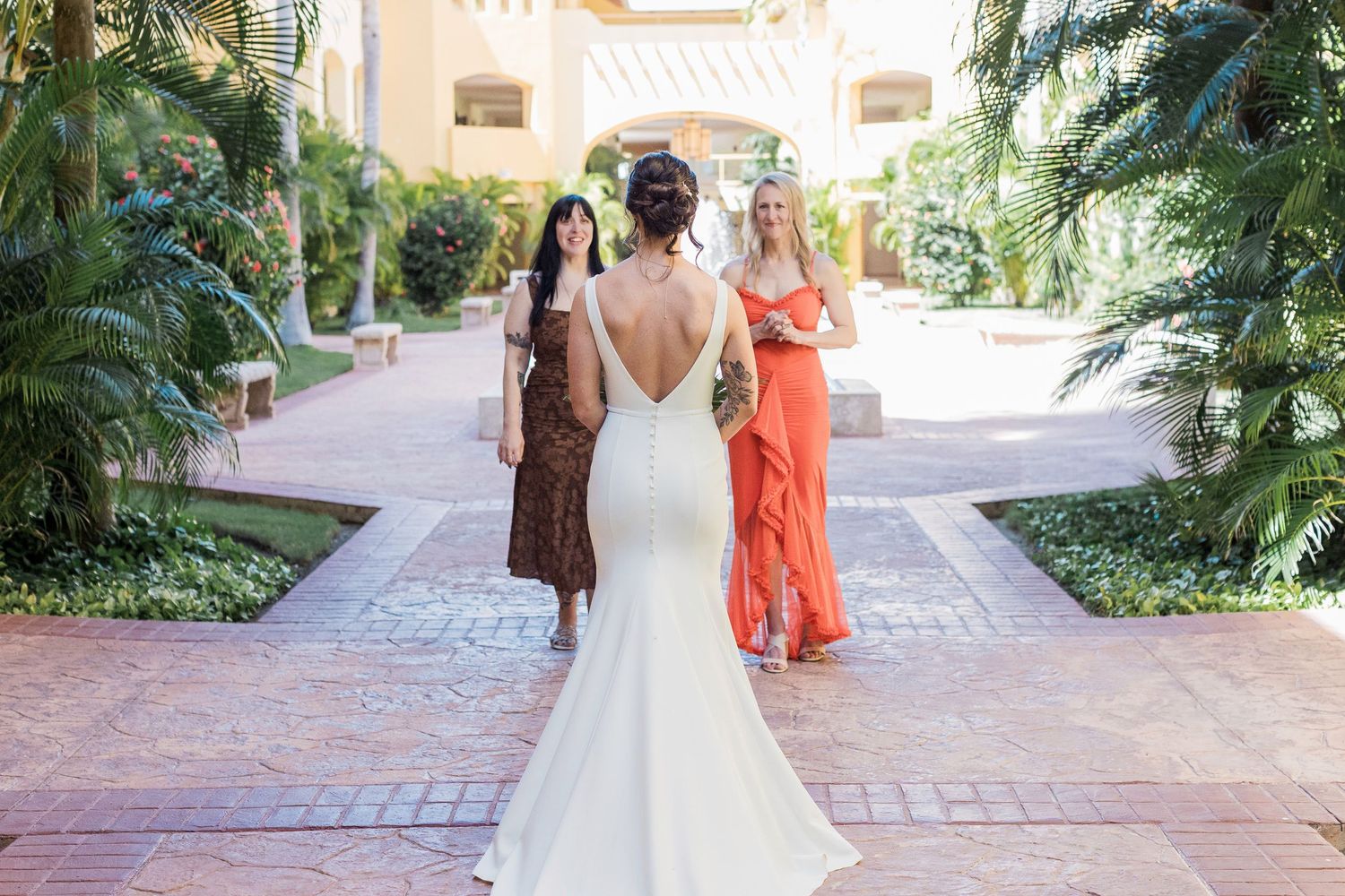 Bride in white backless gown faces two women in colorful dresses at a tropical resort wedding venue.