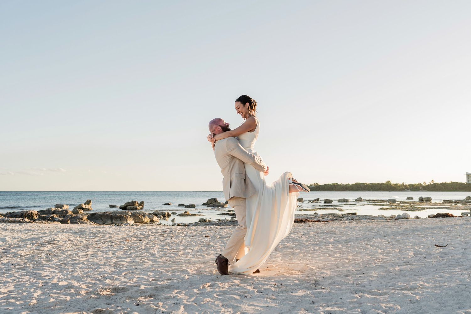 Groom lifting bride in wedding attire on a sandy beach at sunset with calm ocean waters in the background.