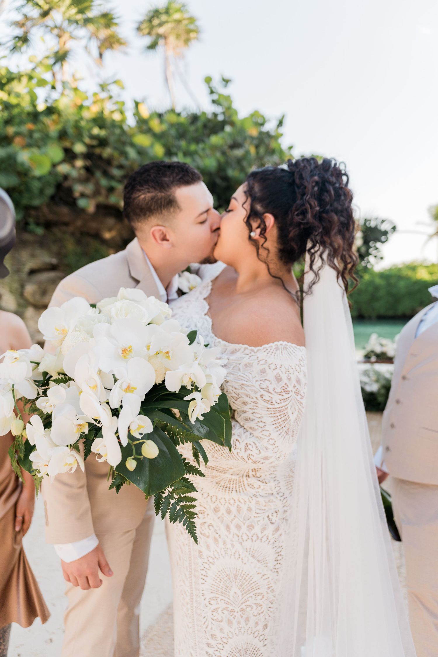 Xcaret Arte Wedding - Bride and groom share a first kiss at outdoor wedding ceremony, bride holding white orchid bouquet, wearing lace gown.
