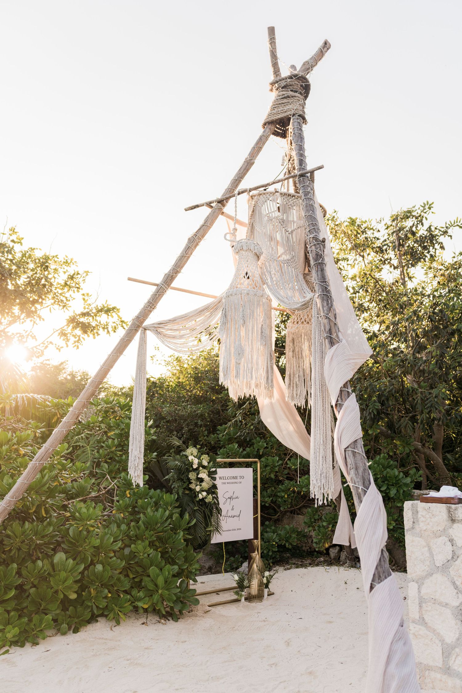 Xcaret Arte Wedding - Boho beach wedding arch made of driftwood poles with macrame and white fabric draping, set on white sand at sunset.