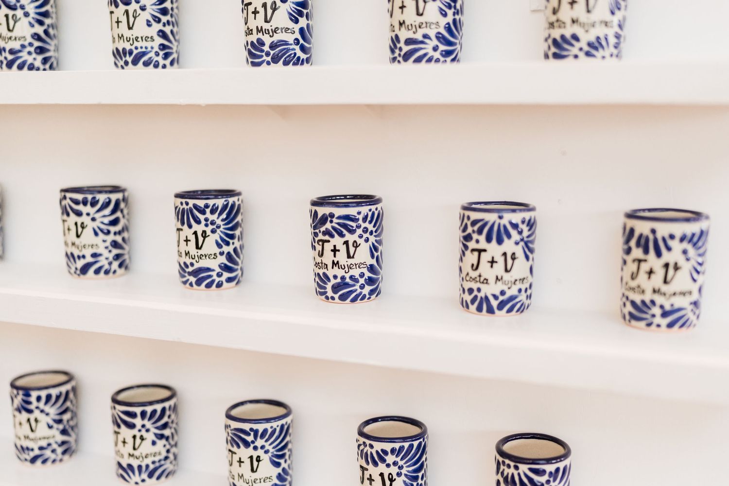 Blue and white hand-painted ceramic shot glasses with monogram designs displayed on white shelves.