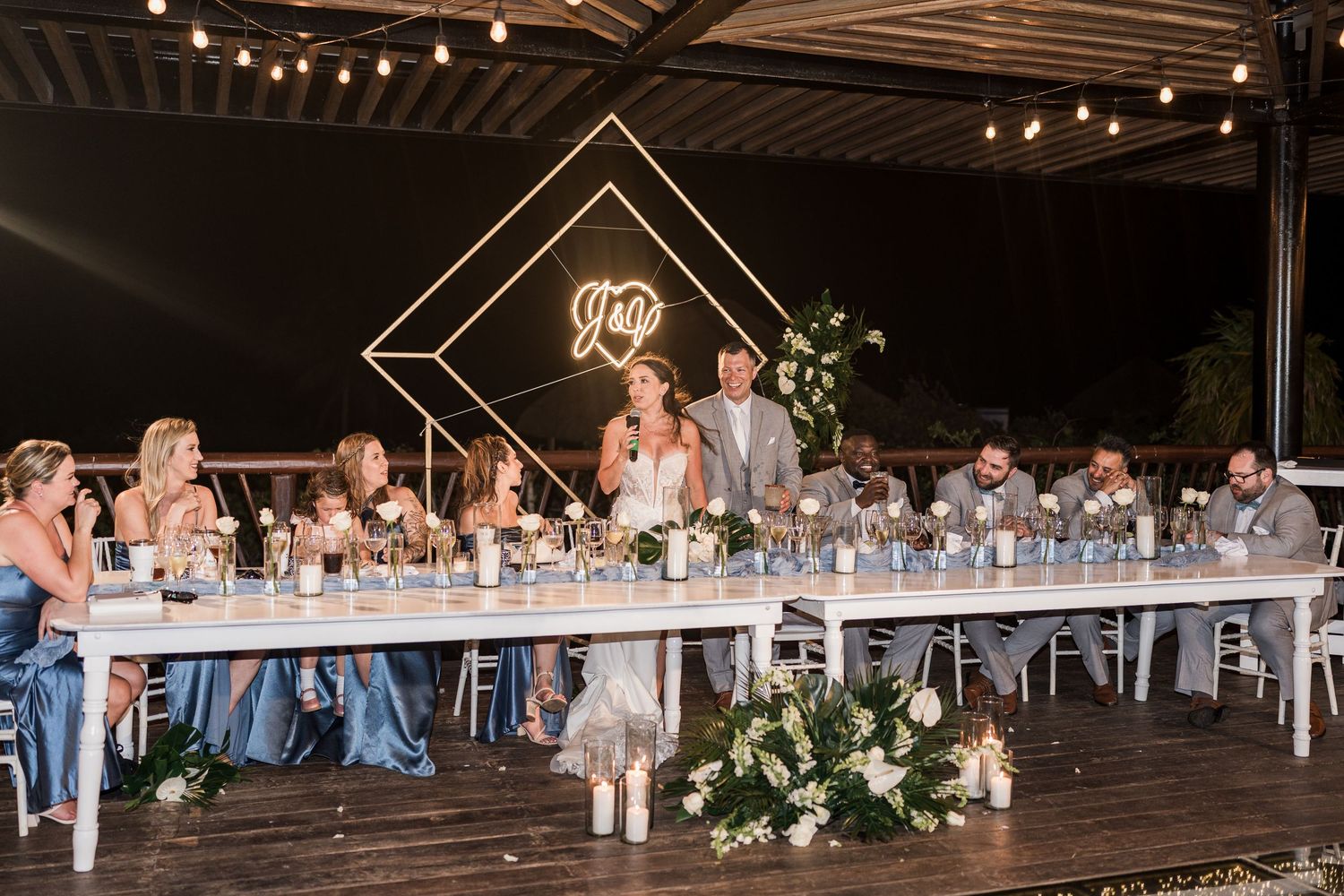 Elegant outdoor wedding reception with string lights, neon monogram sign, and guests seated at a long white table.
