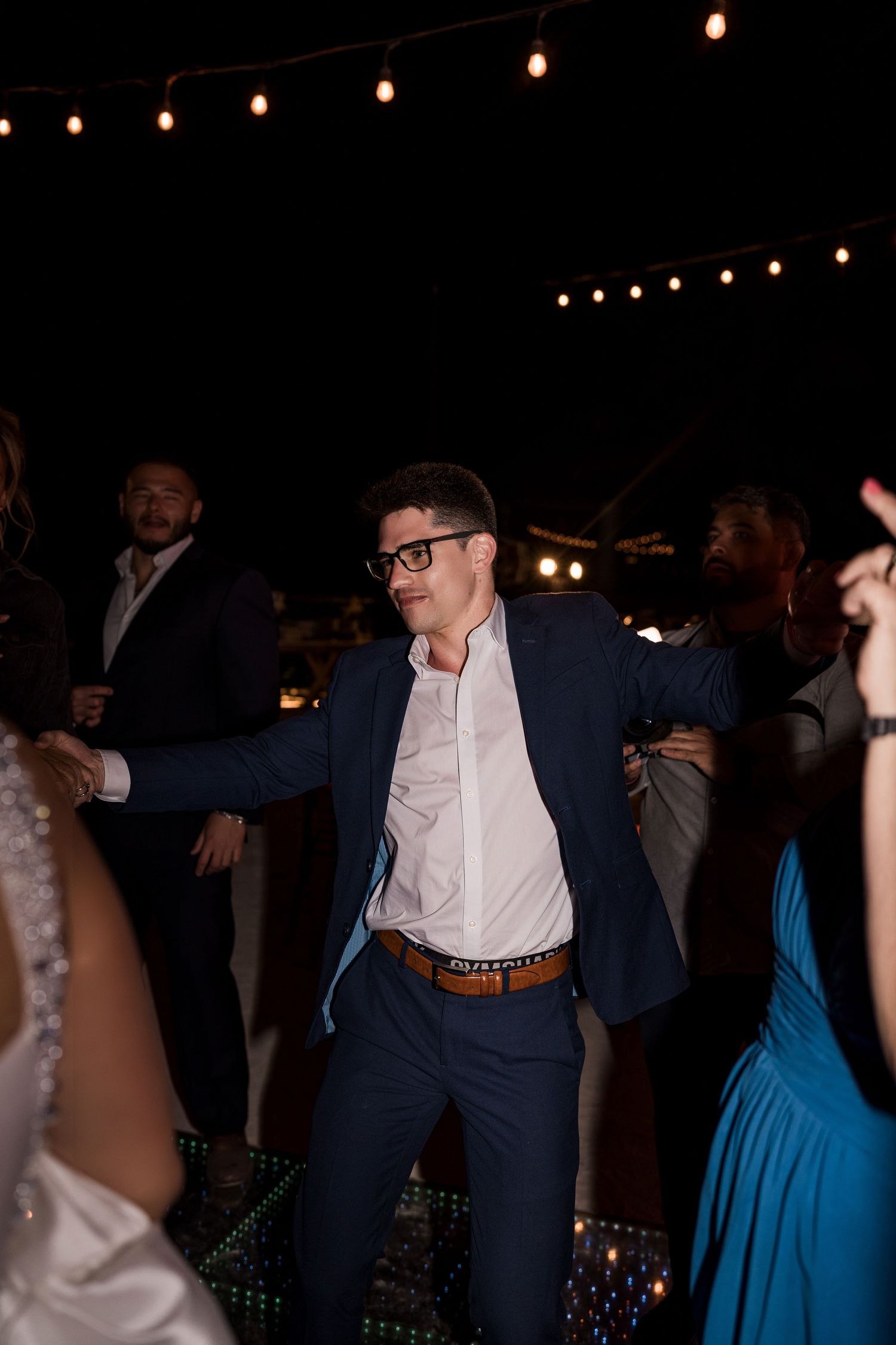 Man in navy suit and glasses dancing energetically at an outdoor evening wedding reception with string lights.