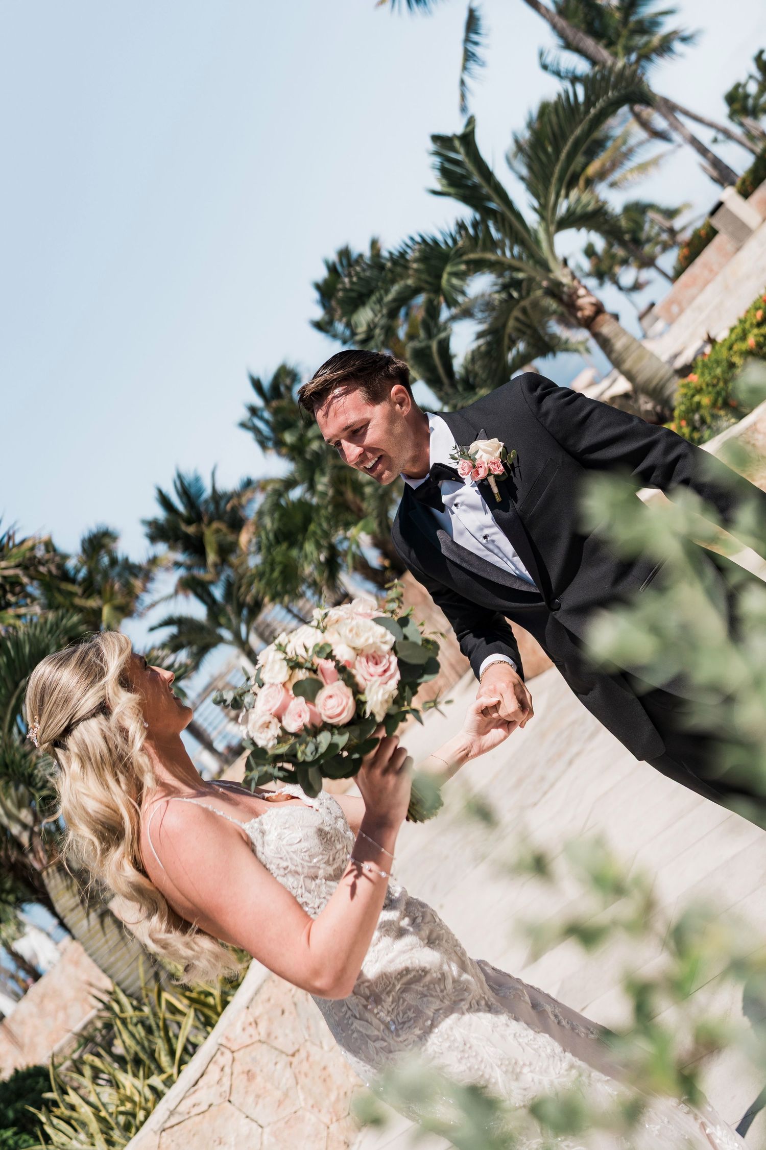 Bride holding pink rose bouquet looks up at groom in black tuxedo, with palm trees and blue sky backdrop.