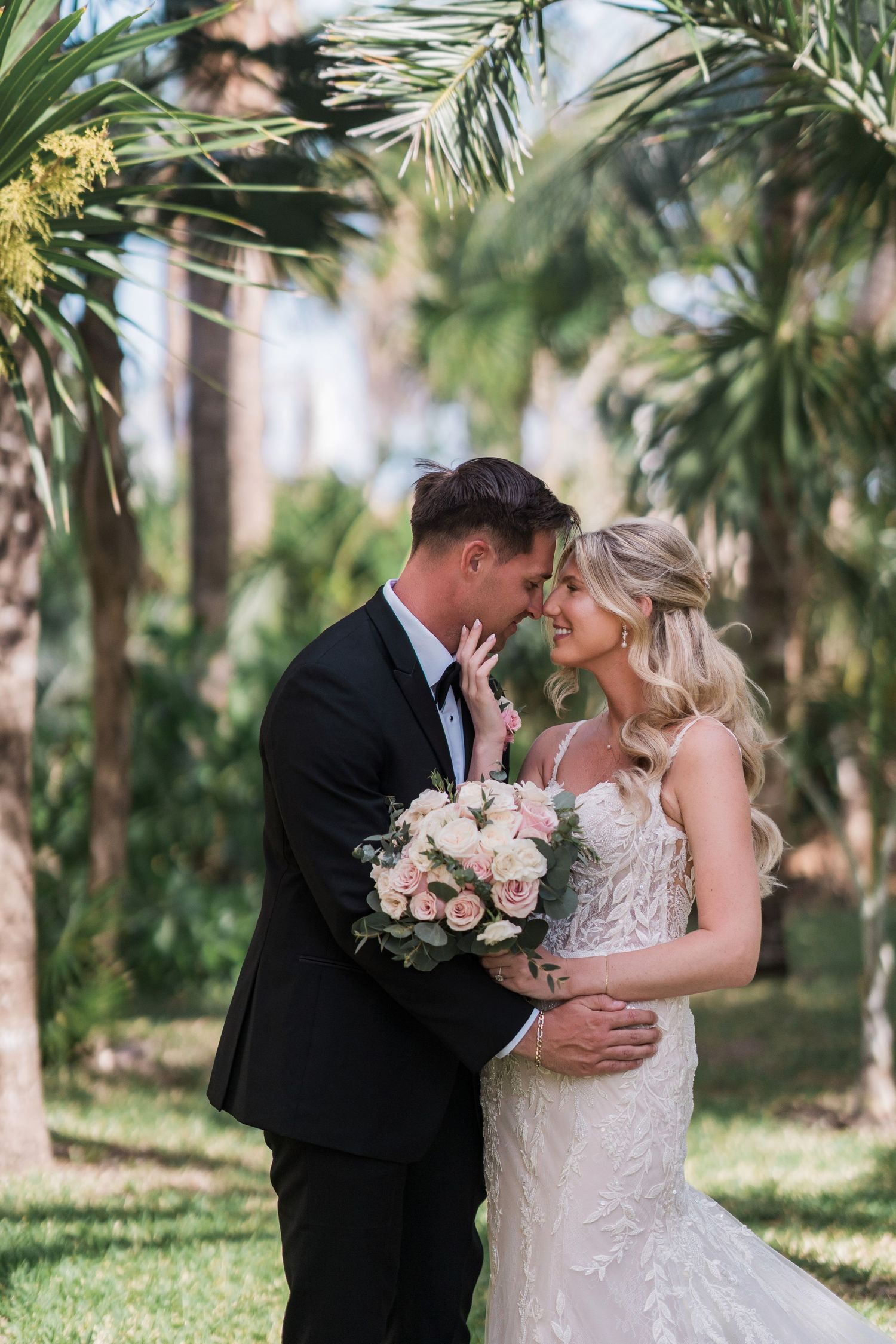 Bride and groom share a romantic moment outdoors surrounded by lush palm trees, bride holding a pink rose bouquet.