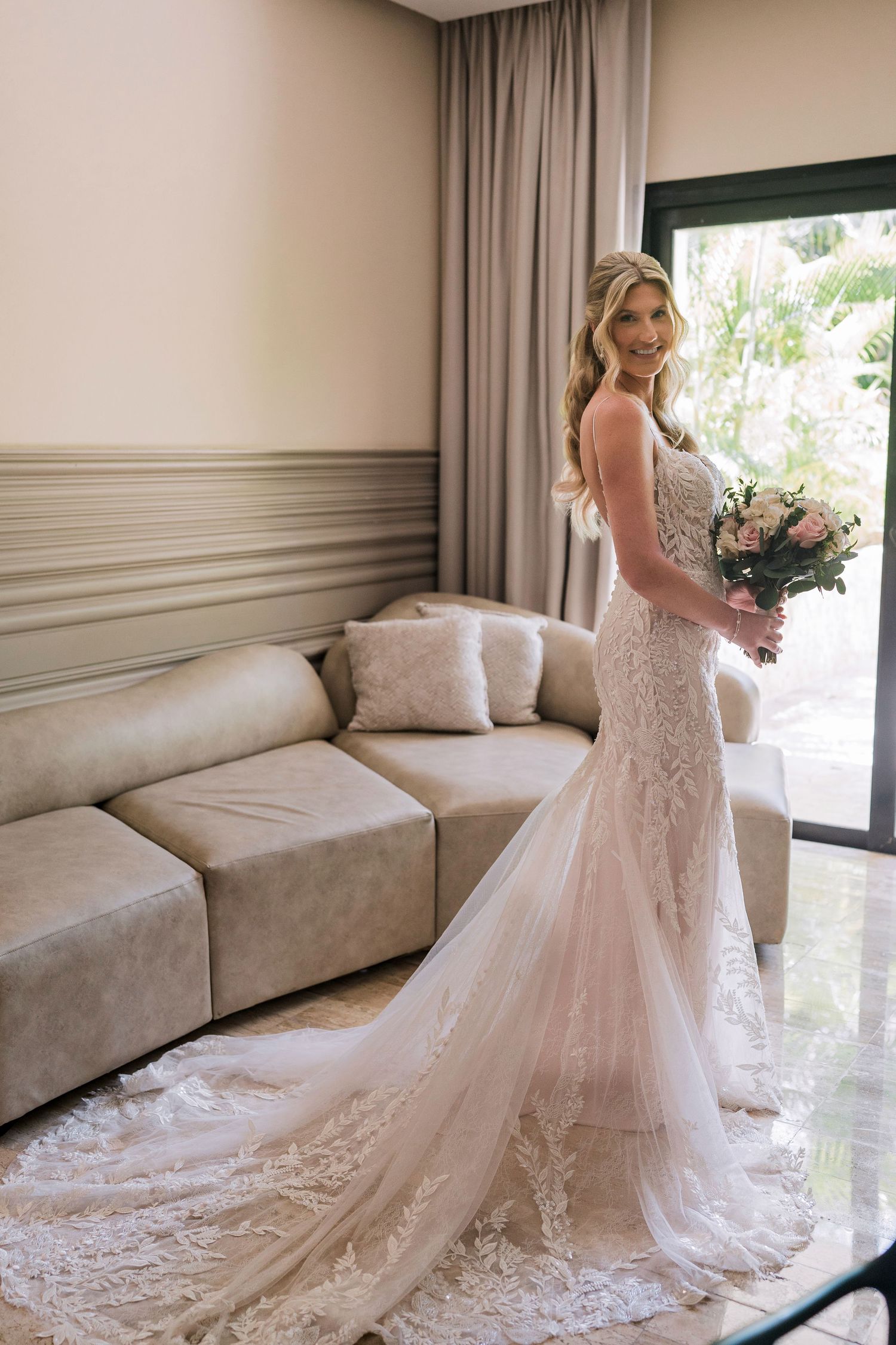 Smiling bride in lace mermaid wedding gown holding bouquet, standing in elegant room with large window.