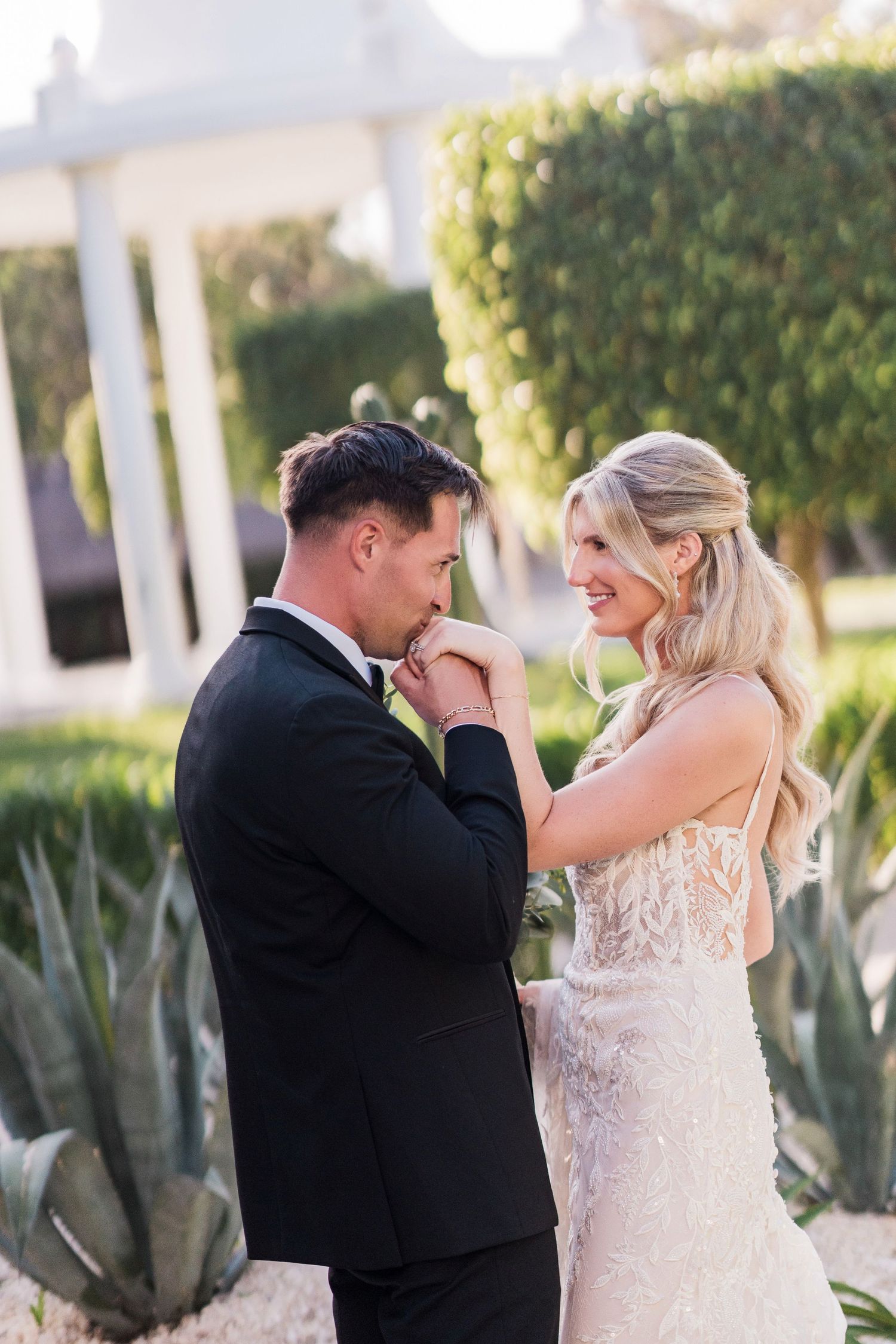 Groom in black tuxedo kissing bride's hand while she smiles in beaded wedding gown at outdoor venue.