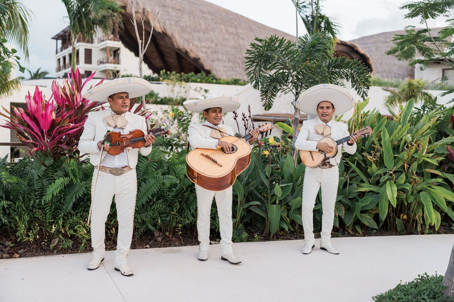 Three mariachi musicians in white suits and sombreros playing instruments at a tropical resort.