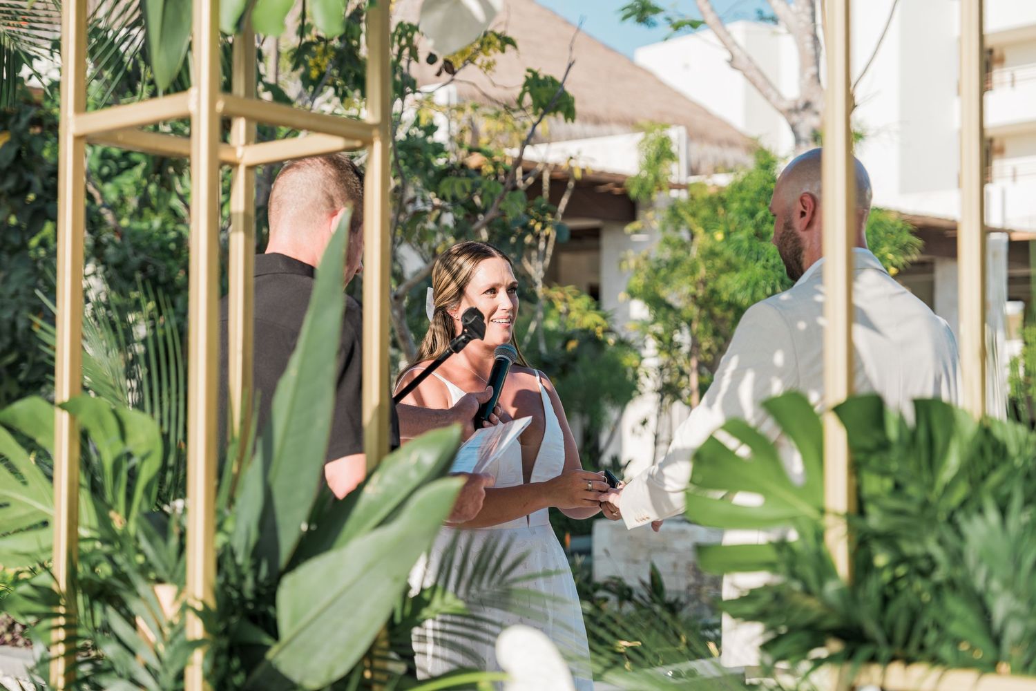 Woman speaking at an outdoor tropical wedding ceremony surrounded by lush greenery and gold geometric arch.