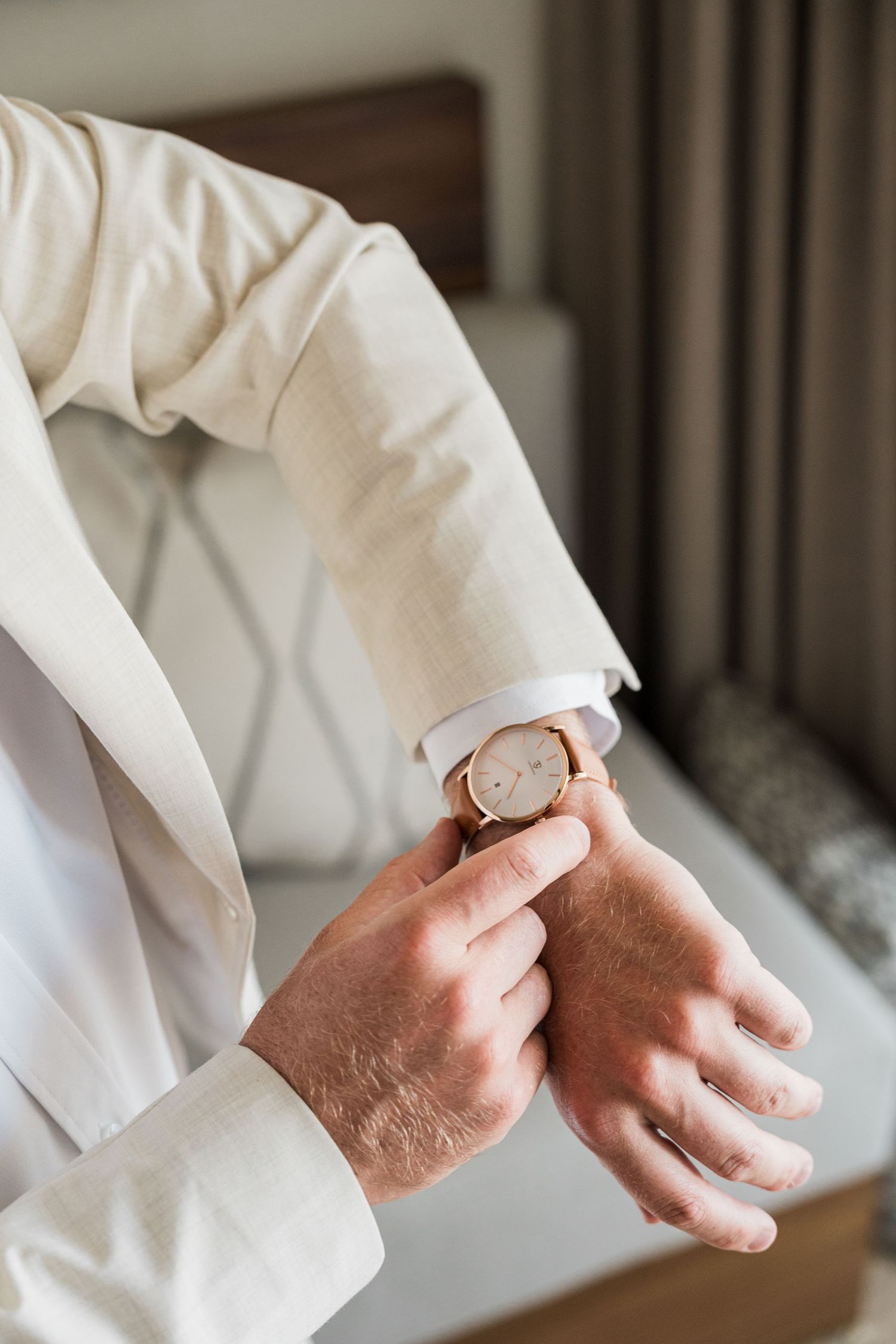 Man in white suit adjusting a gold analog wristwatch, elegant formal attire for a special occasion.