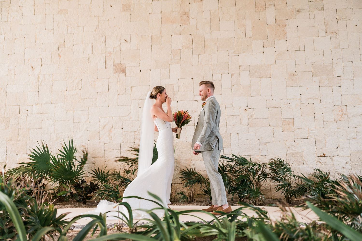 Bride and groom walk hand-in-hand past tropical plants against a stone wall at an outdoor wedding venue.