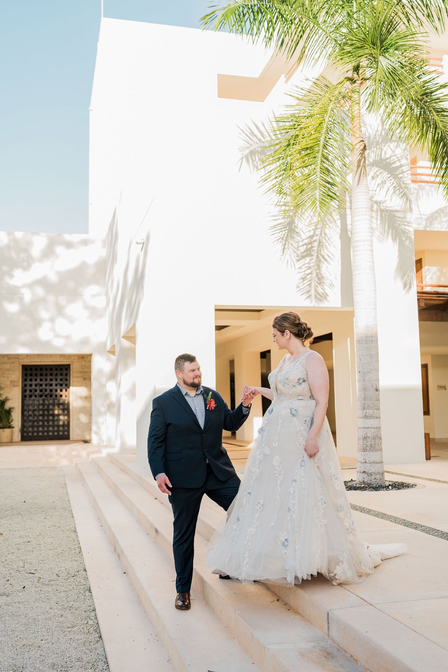 Secrets Akumal Wedding - Bride and groom holding hands on sunlit steps near a palm tree and white building at a destination wedding.