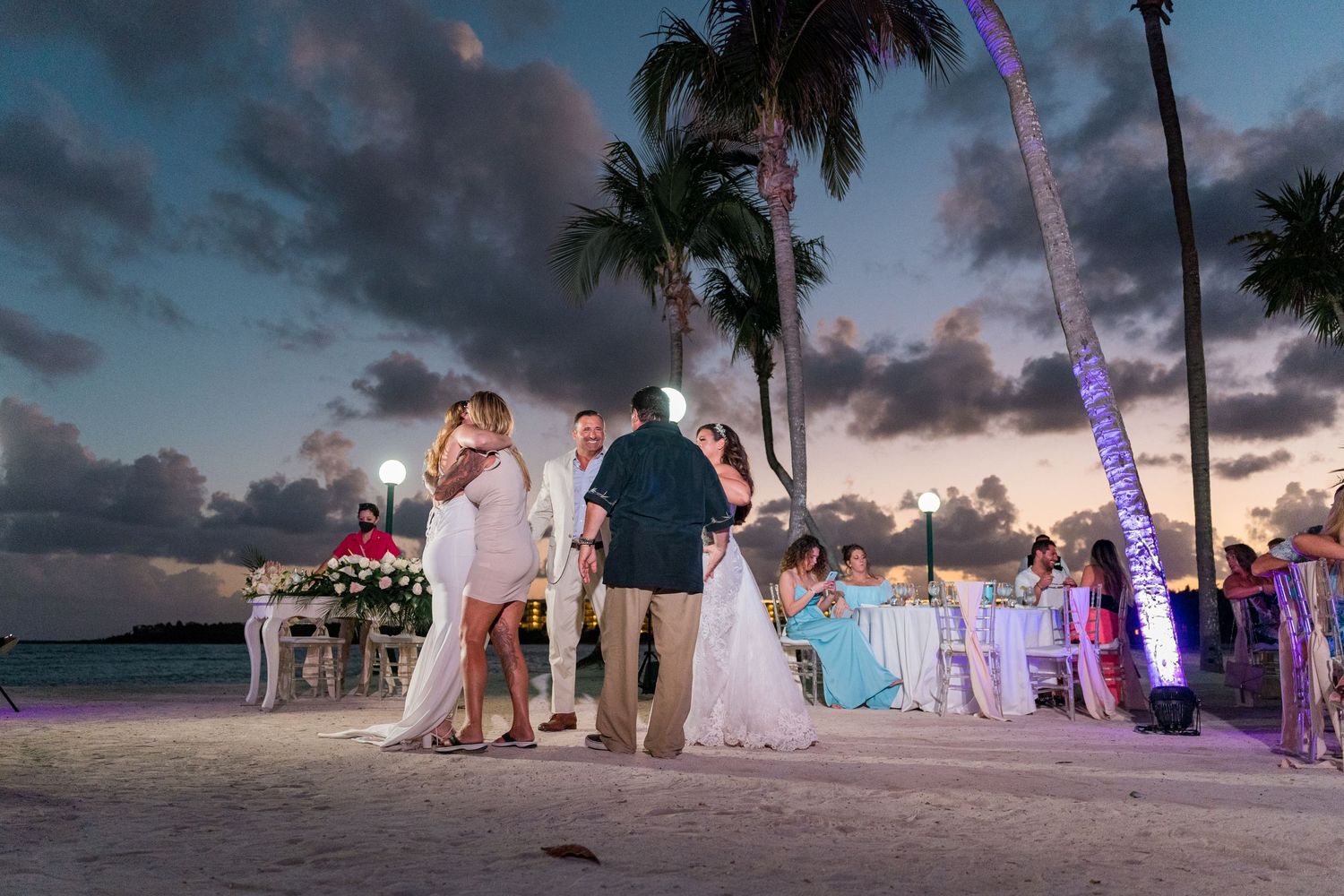 Two couples share romantic dances on sandy beach wedding reception at dusk.