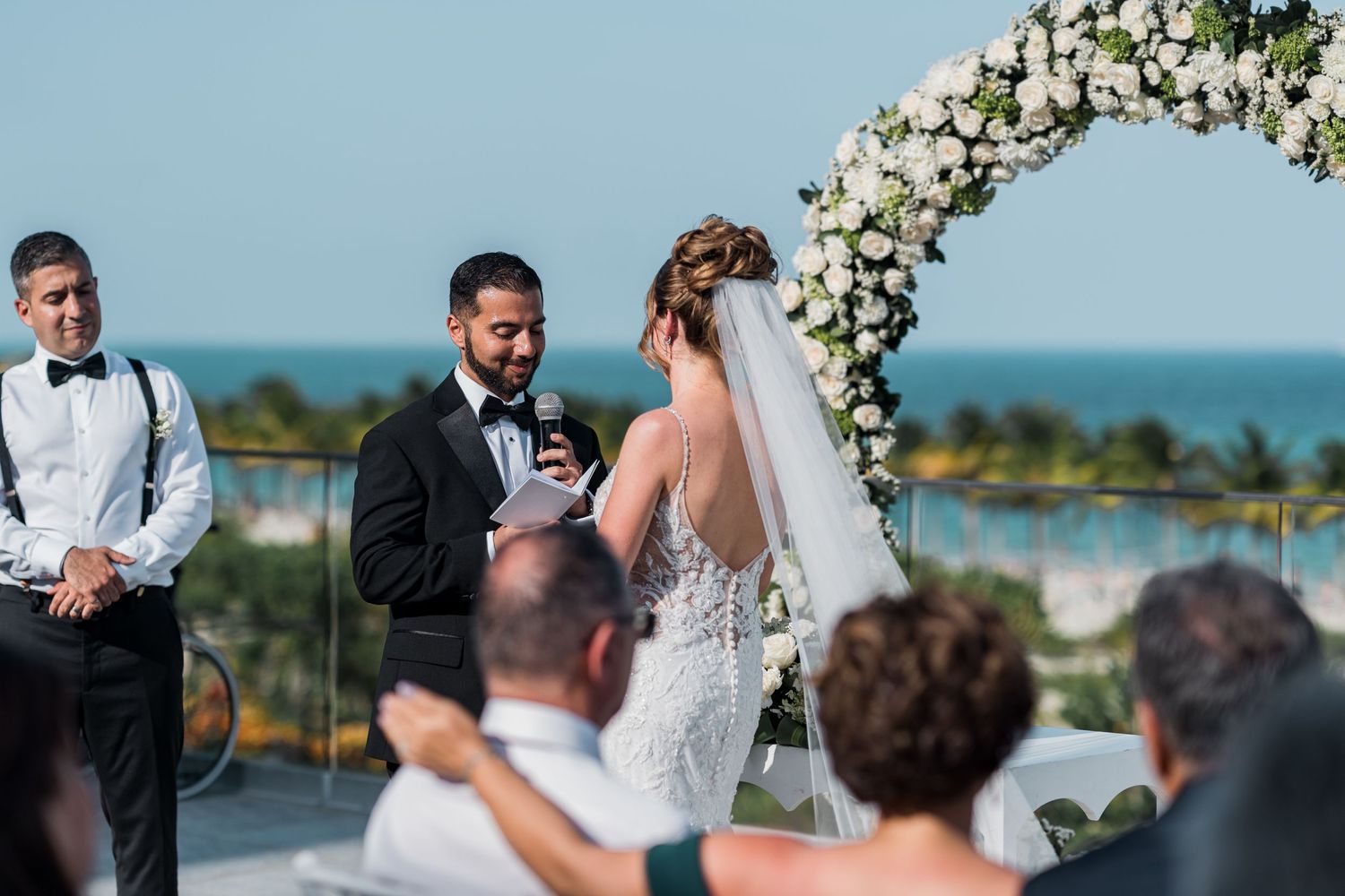 Groom reads vows to bride under floral arch at oceanfront wedding ceremony.