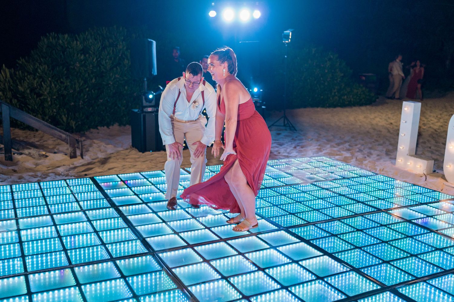 Couple stepping onto illuminated LED dance floor at outdoor beach wedding reception at night.