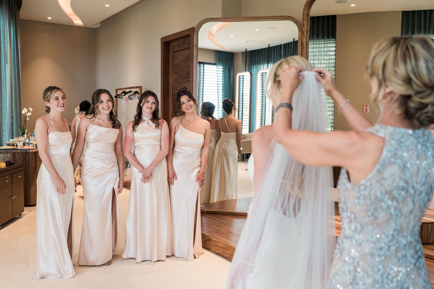 Bridesmaids in champagne gowns watching as mother places veil on bride before wedding ceremony.