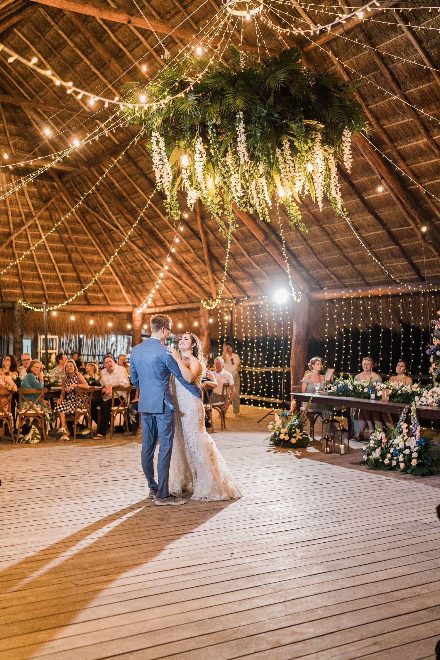 Couple sharing first dance under hanging greenery chandelier and string lights in a palapa venue.