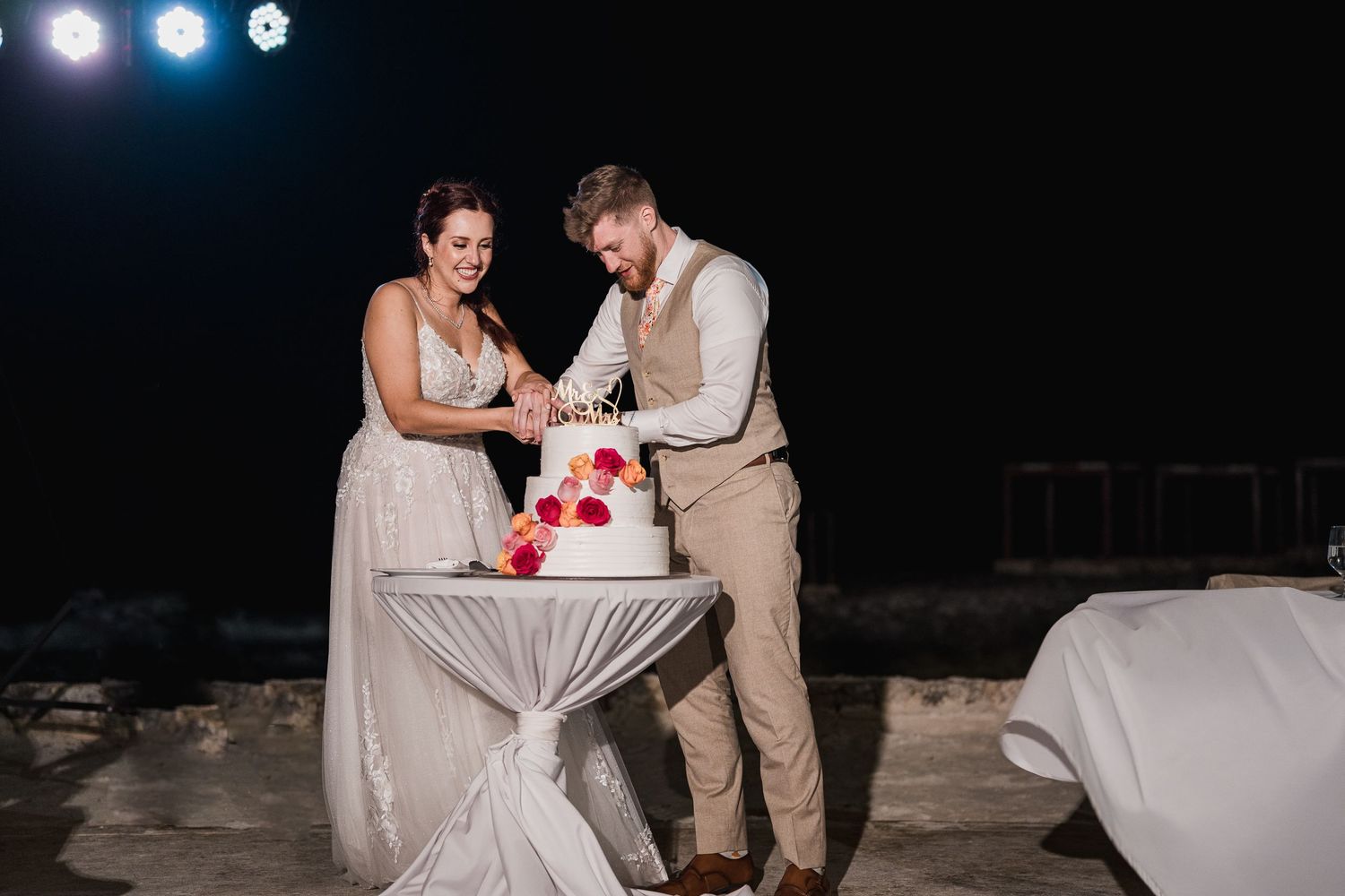 Bride and groom cut their white wedding cake decorated with red flowers at an outdoor nighttime reception.