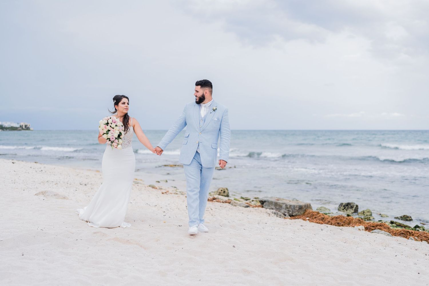 Bride and groom hold hands walking on white sand beach with ocean waves in the background.