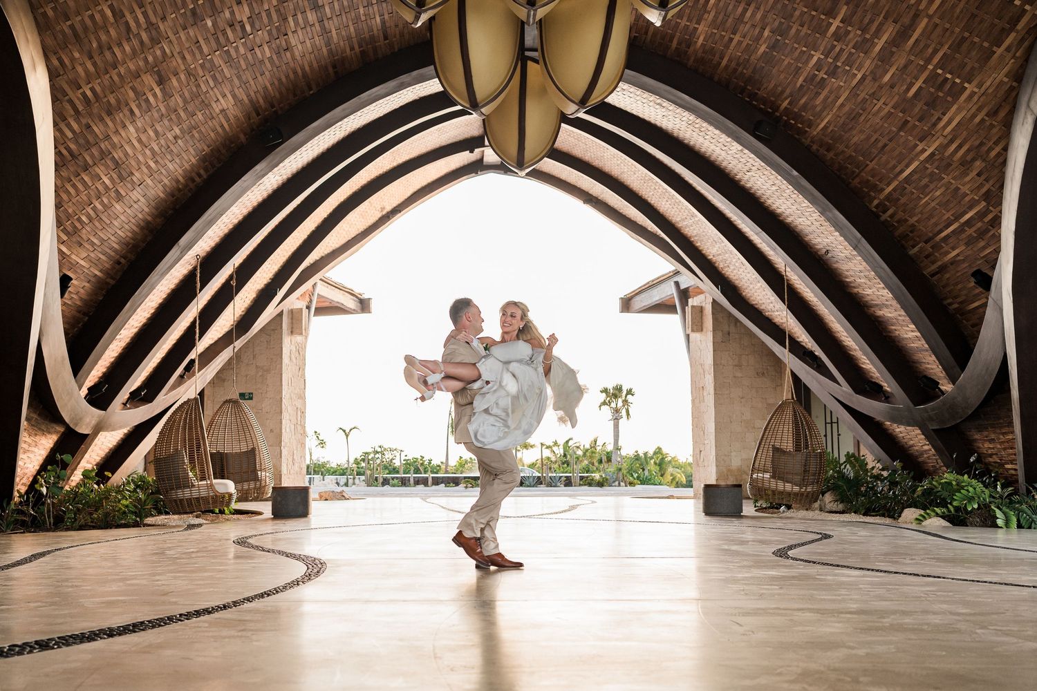 Groom joyfully lifts bride in a sweeping romantic moment beneath an arched tropical resort pavilion.
