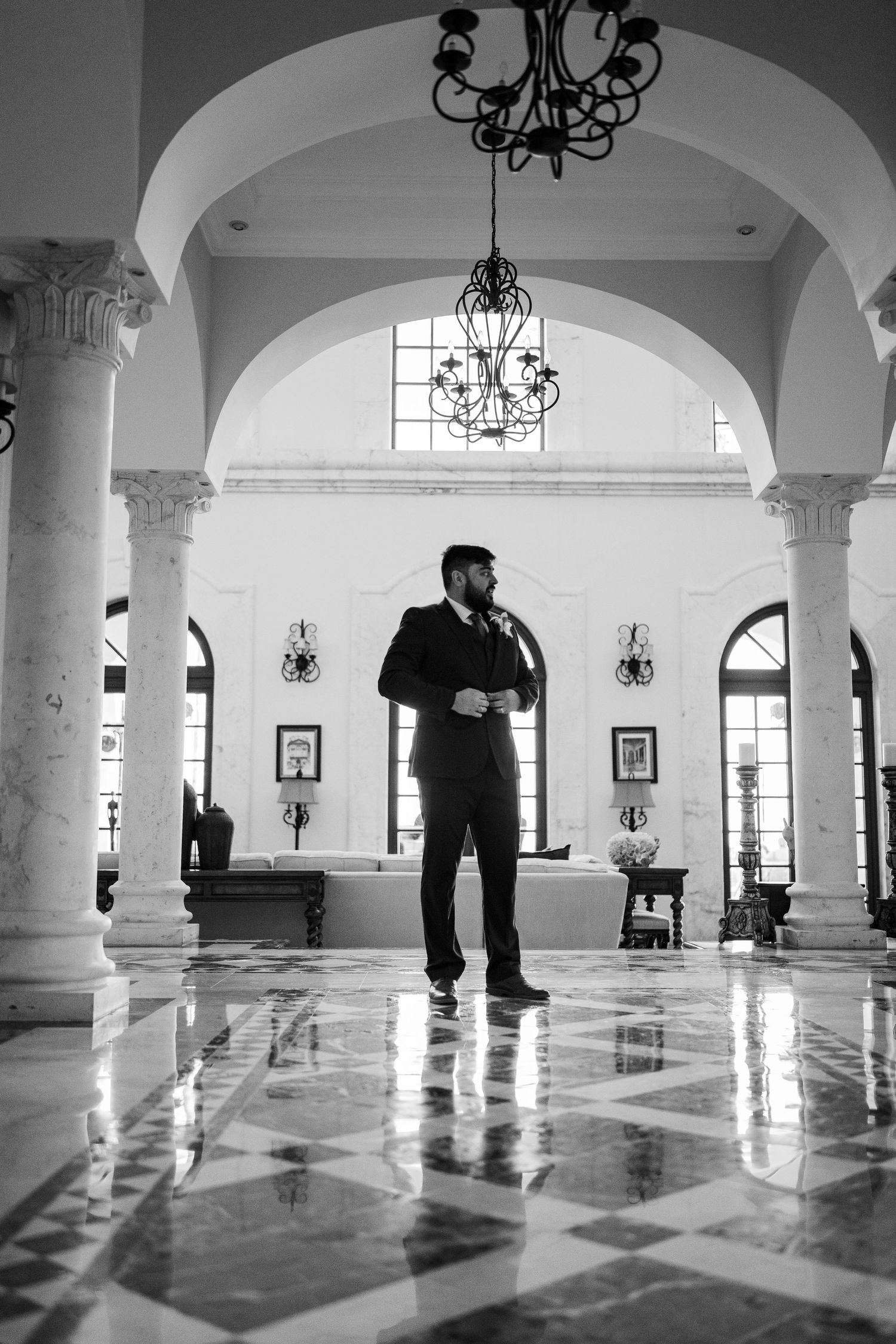 Groom standing confidently in elegant marble lobby with arched columns in black and white photo.