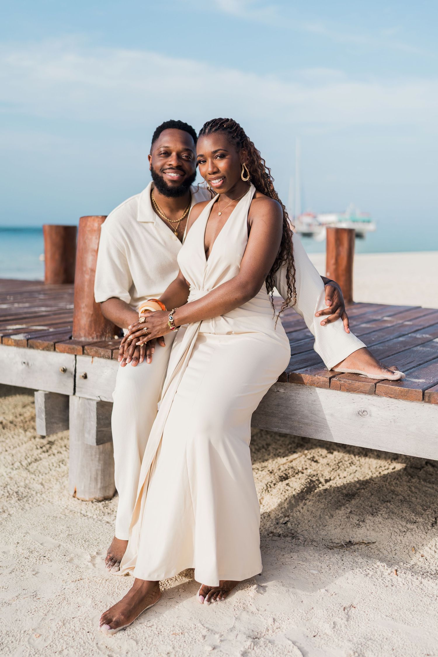A smiling couple dressed in matching cream outfits poses together on a wooden beach dock with turquoise ocean views.