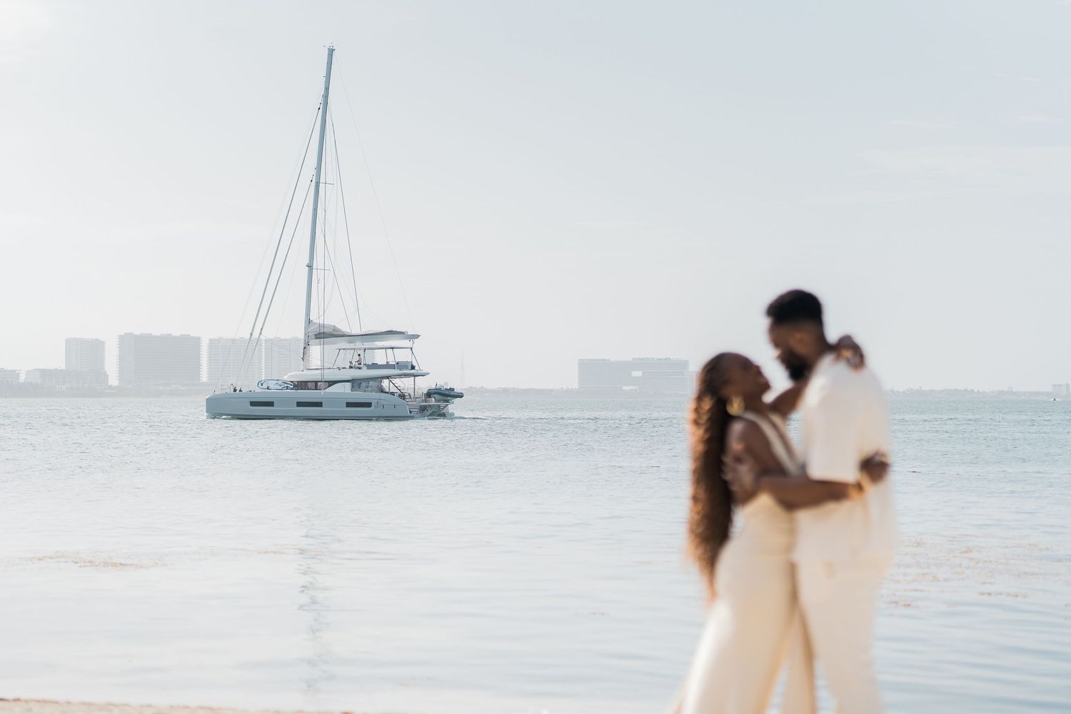 Couple in white attire embracing on a beach with a catamaran sailboat anchored in calm ocean waters behind them.