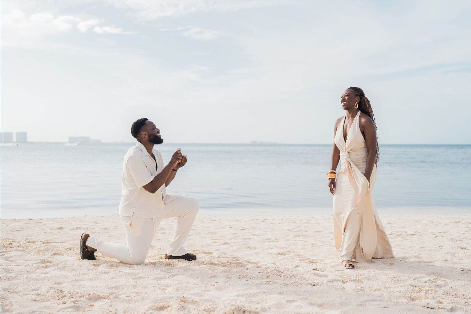 Man proposing on one knee to woman in white dress on sandy beach with ocean backdrop.
