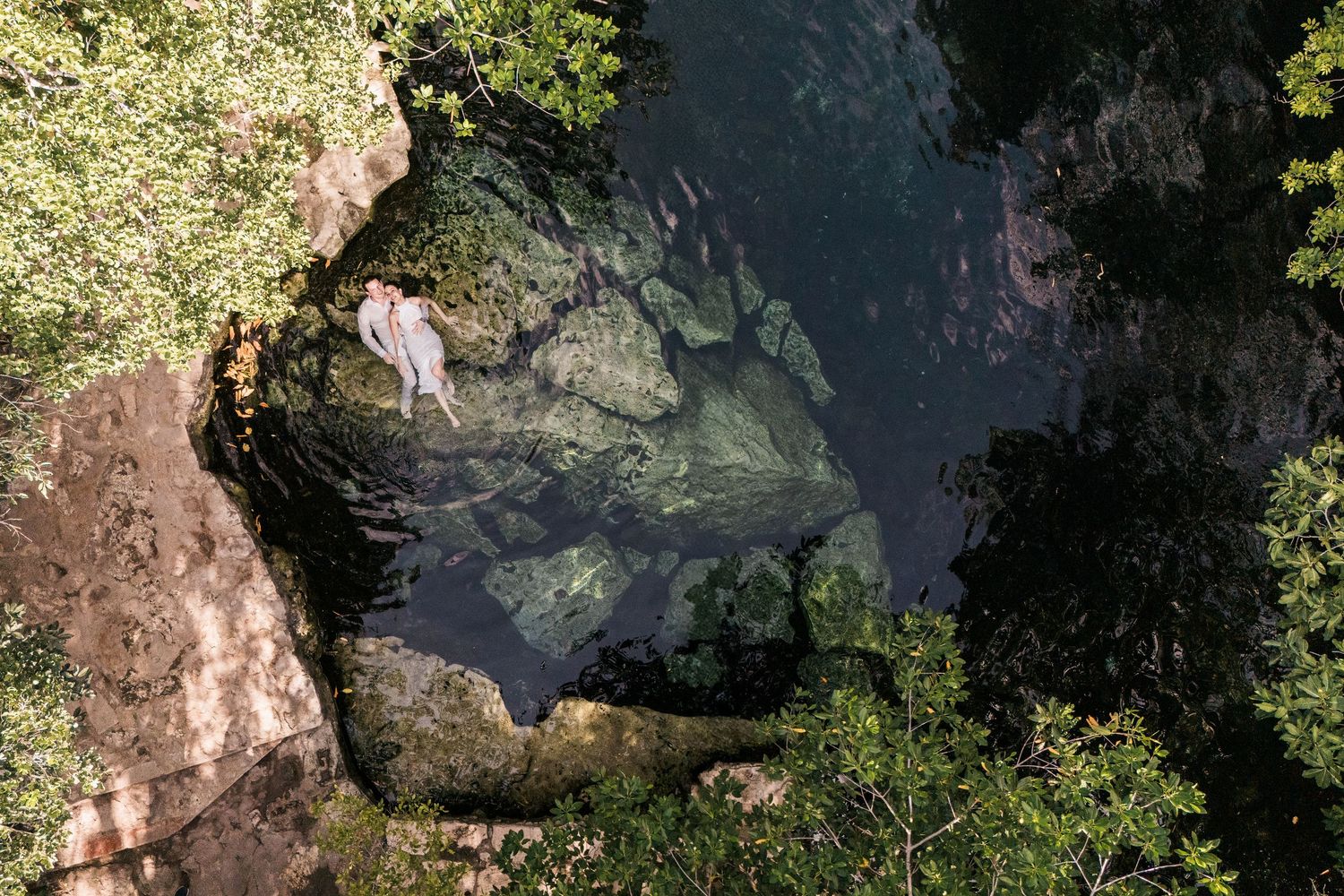 Aerial view of a lush jungle cenote with crystal clear water, rocky edges, and a person standing near the pool.