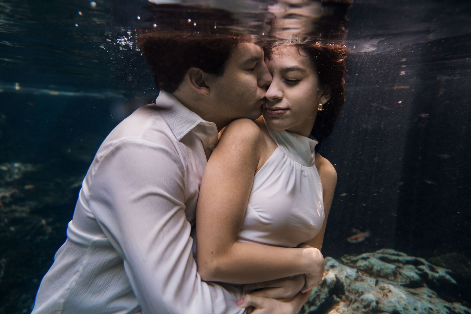 Couple embracing romantically underwater, wearing white formal attire, in a dreamy submerged engagement photo session.