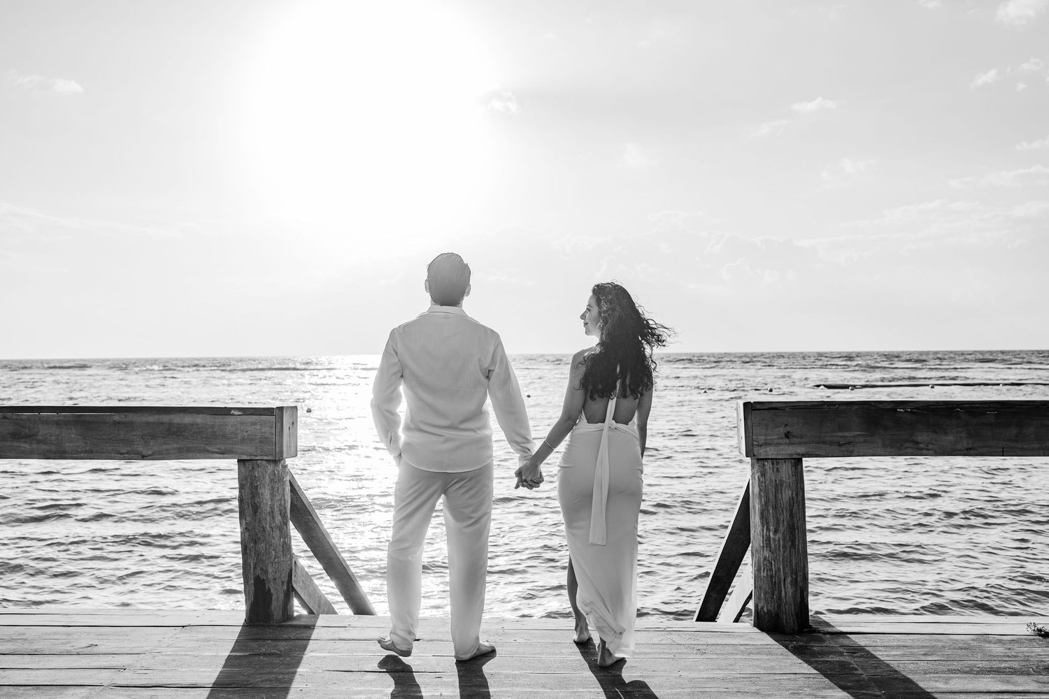 Couple holding hands on a wooden pier at sunset, black and white romantic beach photography.