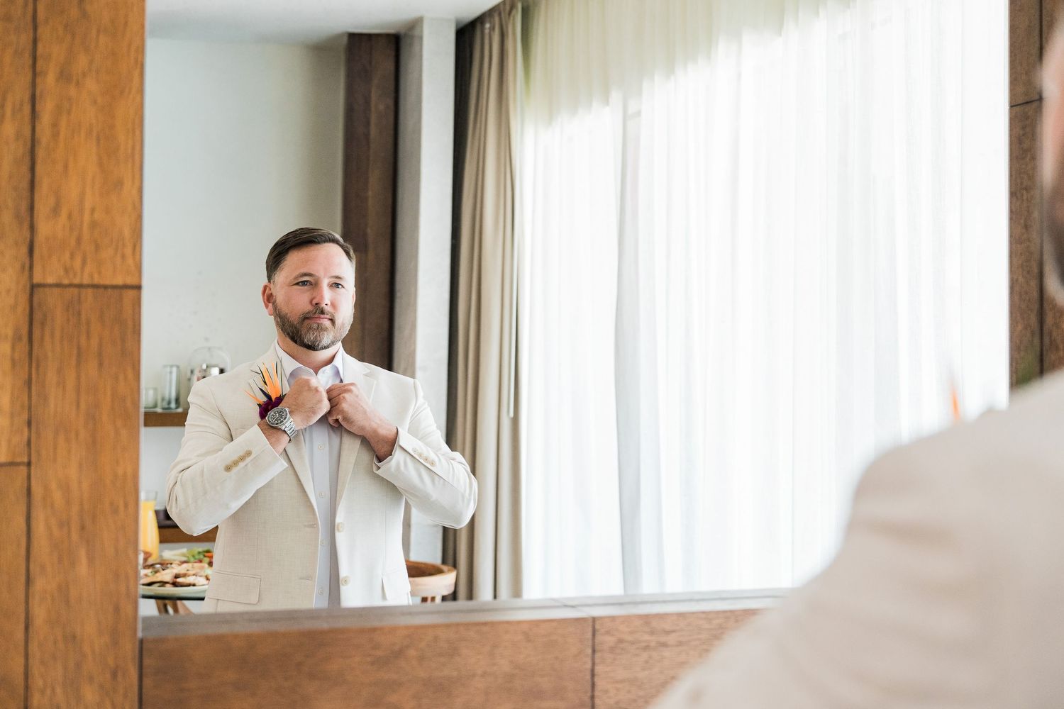 Hotel Xcaret México Wedding  - Man in beige suit adjusting bow tie while looking in mirror in elegant hotel room.
