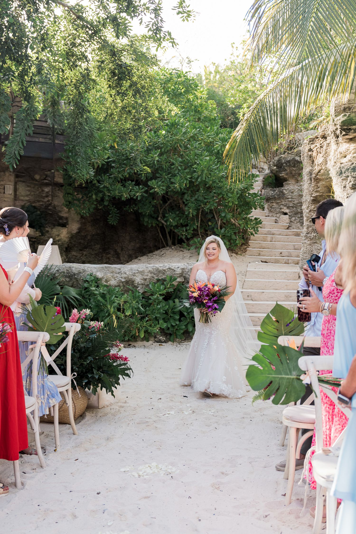 Bride walks down sandy aisle in white gown holding colorful bouquet at outdoor tropical wedding ceremony with lush greenery.
