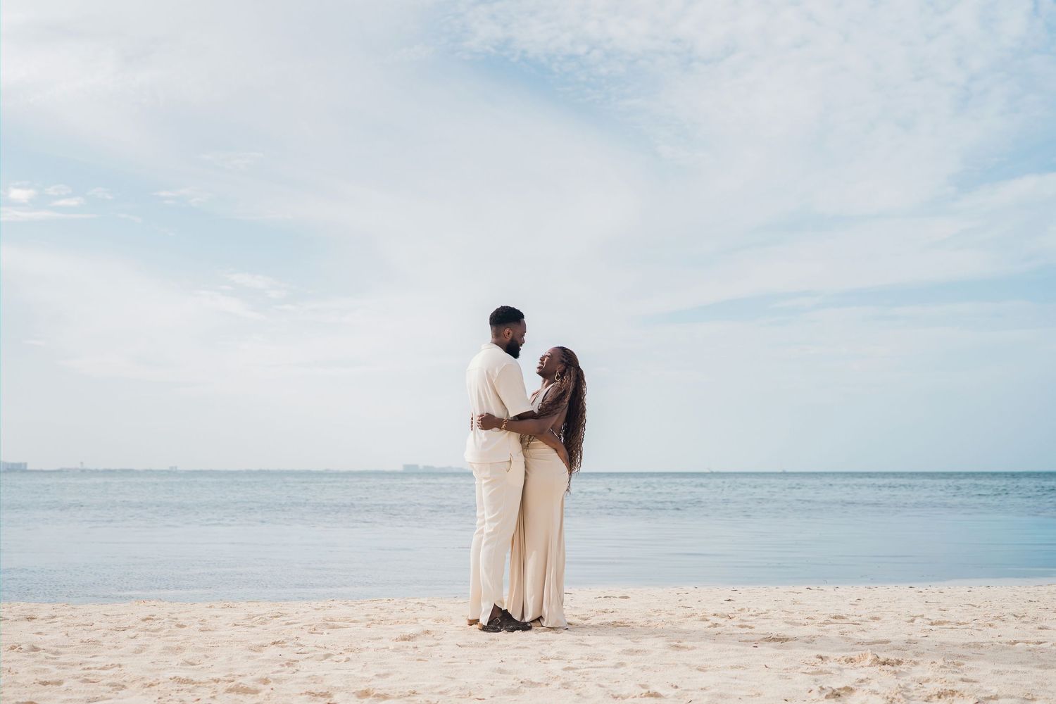 Couple embracing on a sandy beach with calm ocean and cloudy sky in the background.