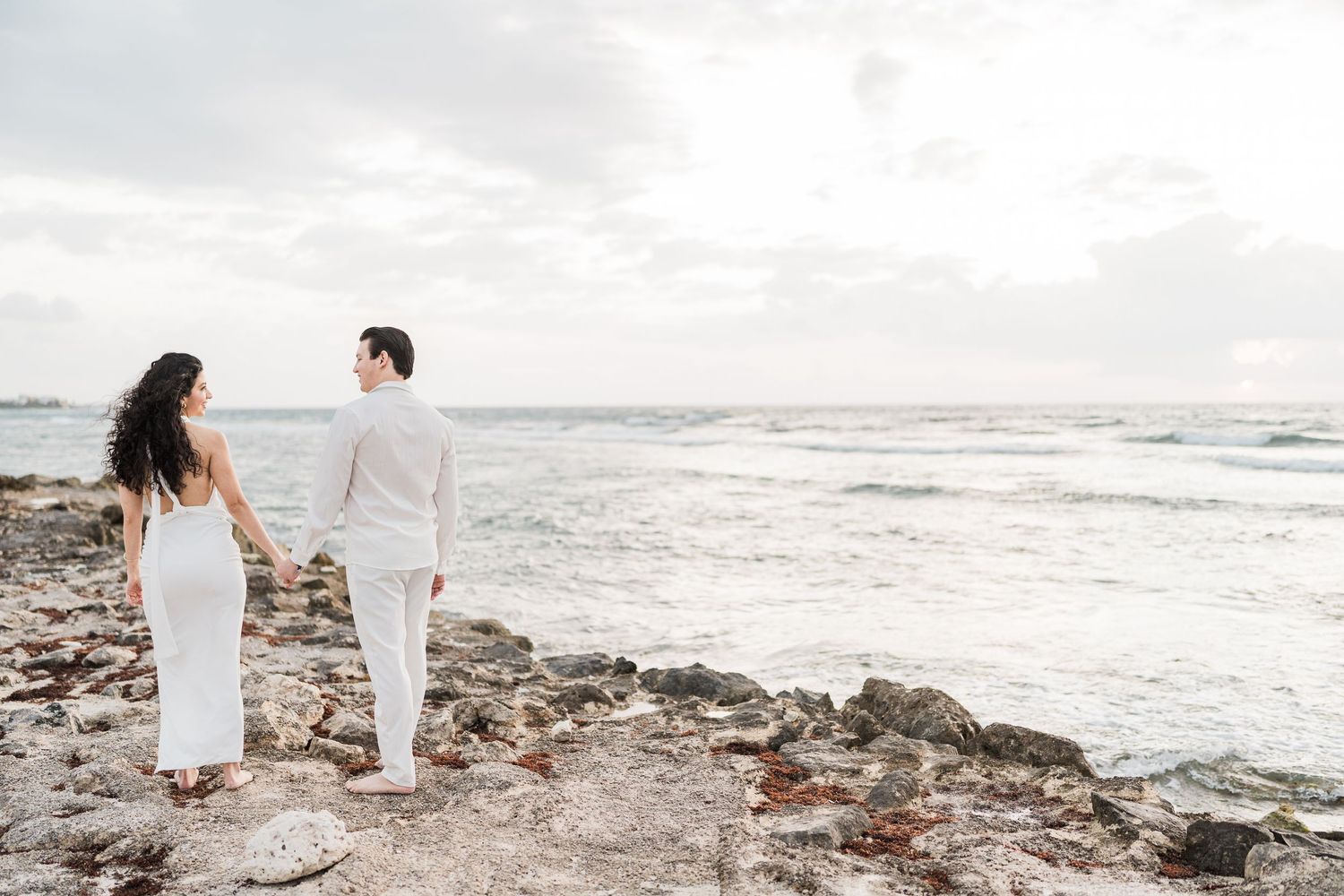 Couple in white holding hands on rocky beach shore with ocean waves and cloudy sky backdrop.