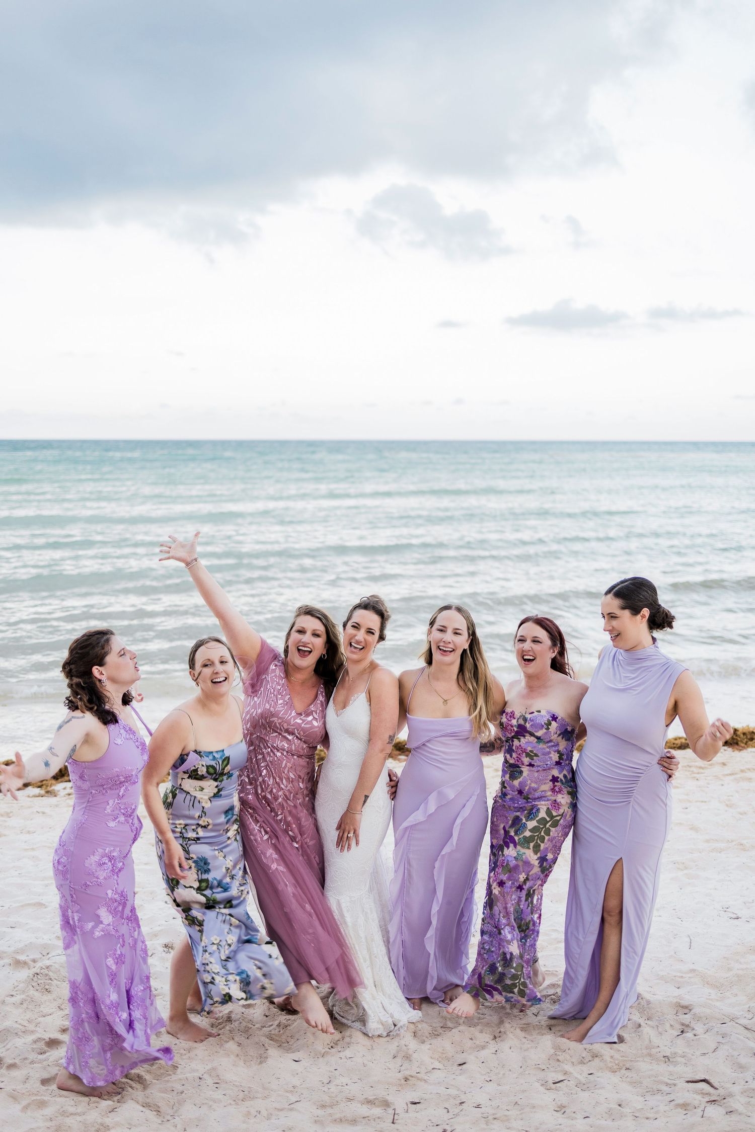 Bride in white gown poses with six bridesmaids in purple dresses on a beach, laughing and celebrating together.